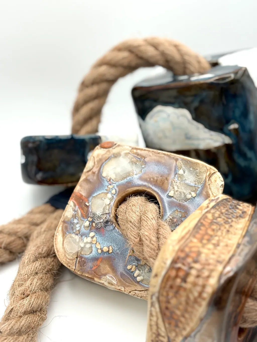 Detail of Bluff/Spit/Drift: a glazed block with a cloud motif threaded on rope, beside a textured brown bead.
