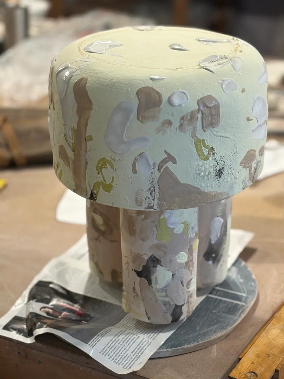 A tall mushroom-shaped form in a pale ochre glaze, drying on newspaper.